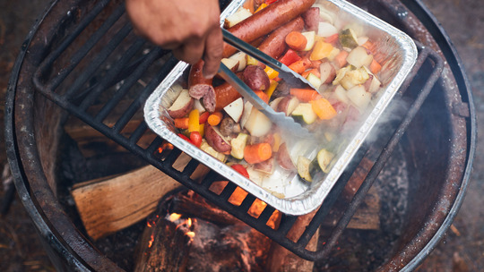 A tin container is cooking a variety of meat and vegetables over a camp fire. 