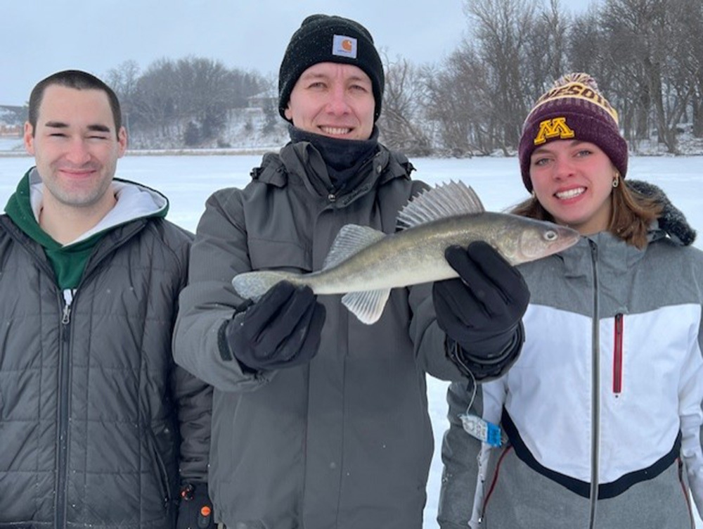 three adults on the ice with center one holding a walleye