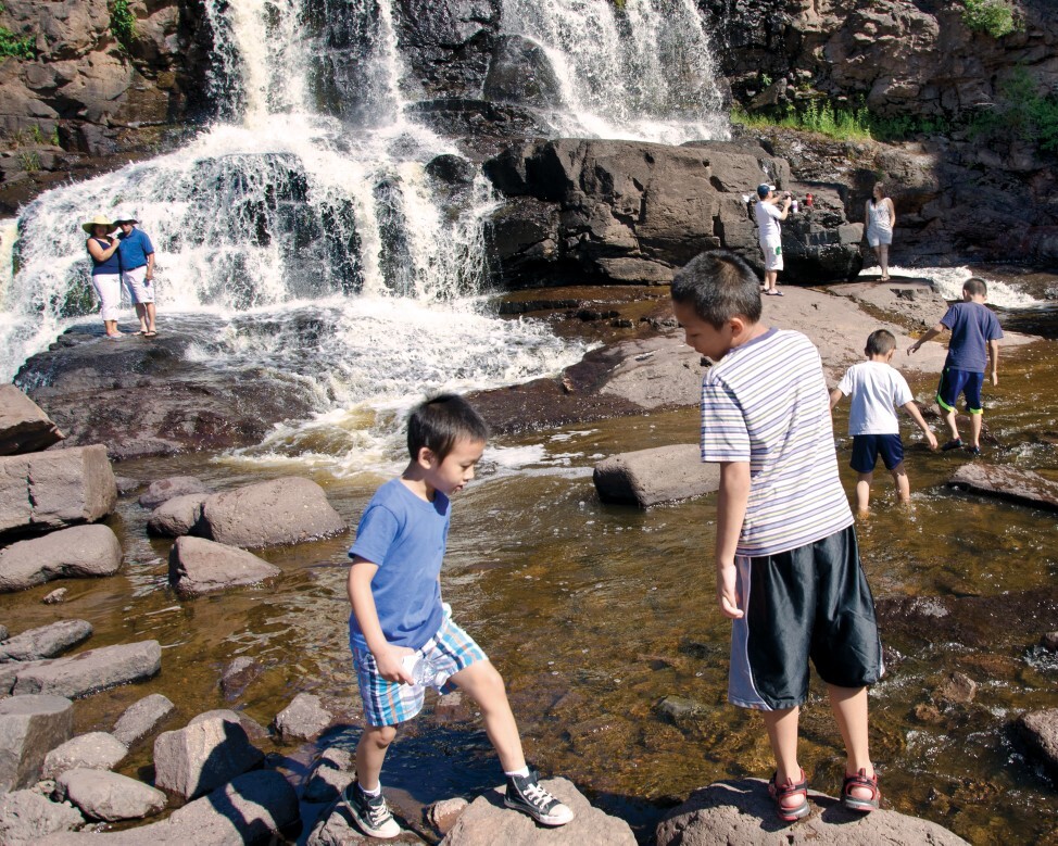 Kids and adults play in the water below a waterfall.
