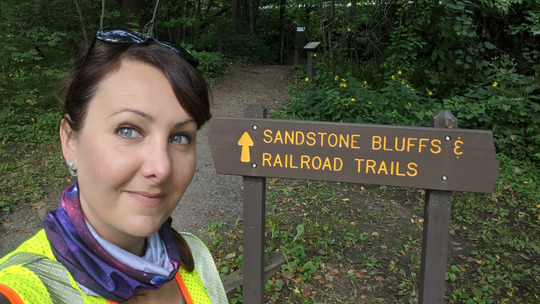 Amy smiles in front of a brown state park that points to Sandstone Bluffs Railroad trails. 