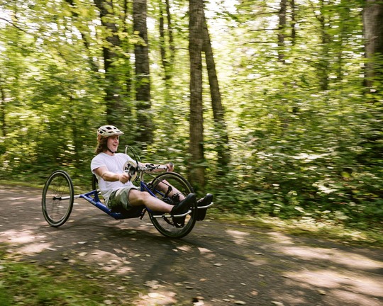 A young man smiles broadly while using a handcycle bike on a paved trail.