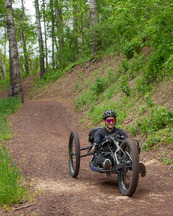 A person smiles while using a handcycle down a curing dirt path. 