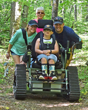 A family smiles and crowds around a child sitting in an all terrain track chair in a forest. 
