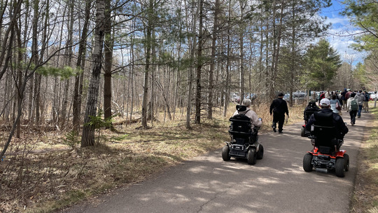 Two electric wheelchair users follow behind a group of people walking on a paved trail.