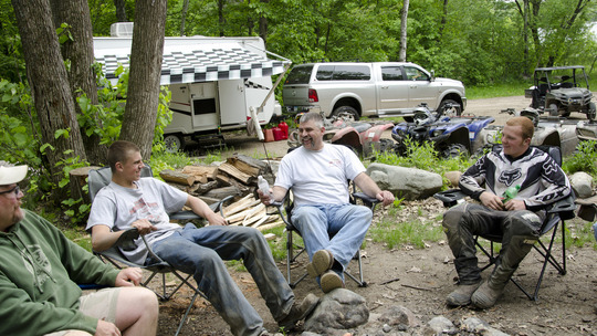 A group of campers gather around a fire with OHVs and a trailer in the background. 