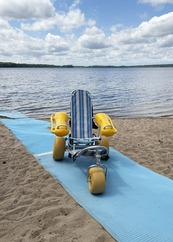 A blue and white striped beach chair sits on a mat that stretches across a sandy beach to the water. 