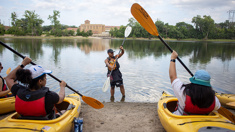Devin Brown stands with a paddle in front of a river showing a group of people how to kayak. 