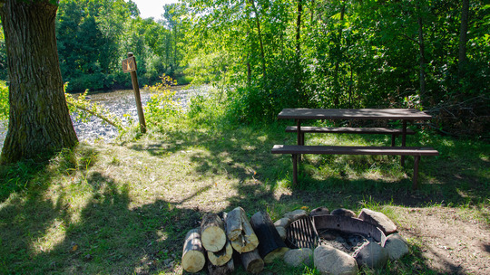 A campsite has a picnic table, fire ring, and river in the background. 