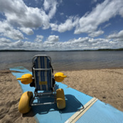 An accessible beach chair sits atop a beach mat that extends down a sandy shoreline. 