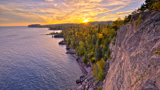 A wide landscape of the Lake Superior shoreline, bordered by rocky cliffs and deep green pines.