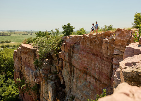Two people stand on the edge of a towering, rusty red cliffside.
