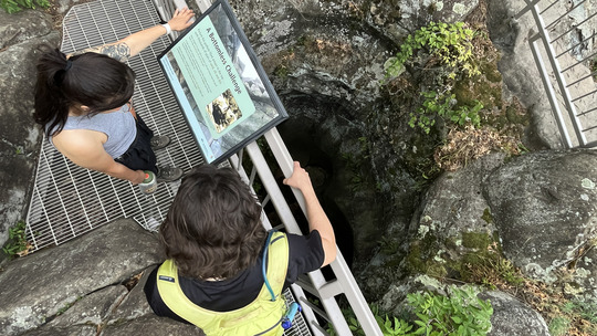 Two people read a sign next to a deep cavern in the rock that extends down into the darkness.