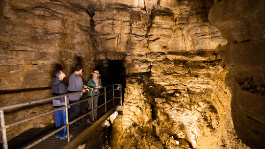 Two teens stand with a park ranger. They look around at an underground cave with tall rock walls.