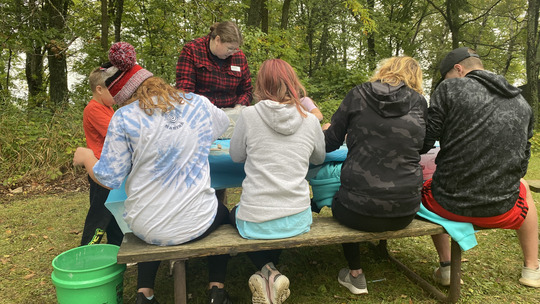A group of people crowd around a picnic table while looking at small pieces of stone and dirt.