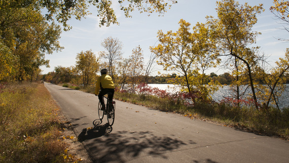 A person bikes along a smooth paved trail stretching into a fall landscape.