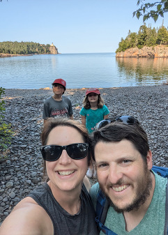 Two adults take a selfie with two children behind them. In the background is the North Shore and Split Rock Lighthouse. 