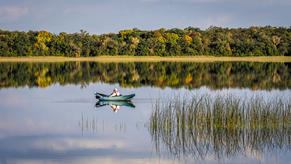 A kayaker pauses on a calm lake surrounded by brilliantly colored trees in fall. 