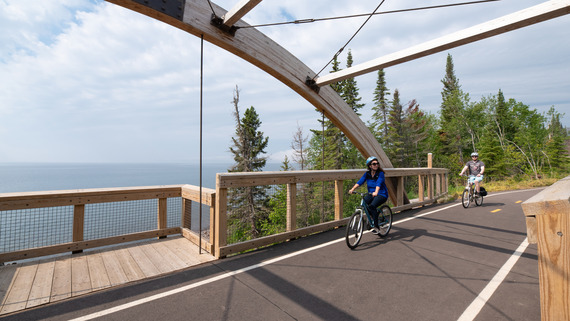 Two people bike along a paved path next to the shoreline of Lake Superior. 