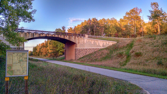 A paved path passes under a small stone bridge. The whole area is forested with bright fall foliage. 