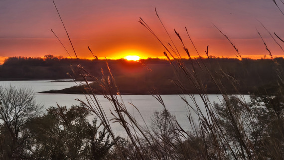 A brilliant red sunset over a calm lake with tallgrass in the foreground. 