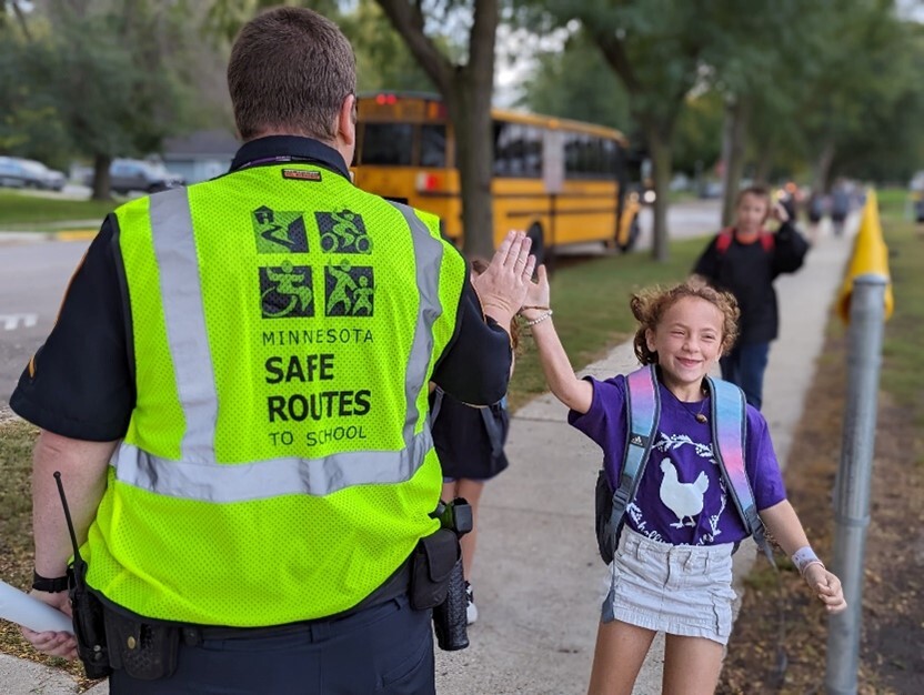 A girl walking down the sidewalk giving a high five to an adult safety patrol. 