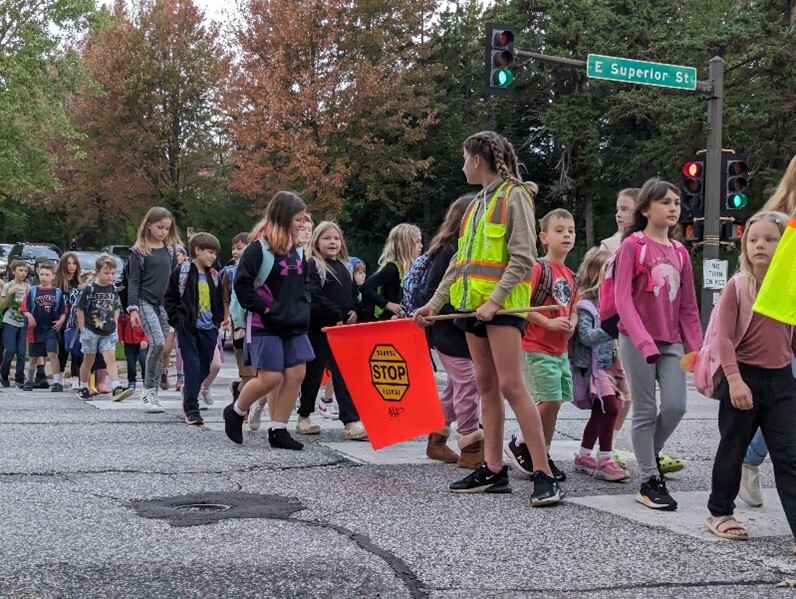 A group of students are walking to school; while the crossing guard is standing in the street holding the stop flag.