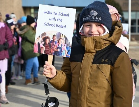 Student holding a walk to school with the school board sign. In from of the school with students in background.