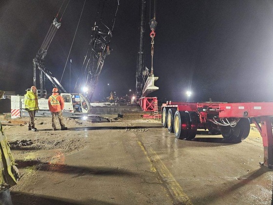 Photo of bridge beam moving to Fourth Street Northwest bridge with construction inspectors standing nearby on a rainy night.