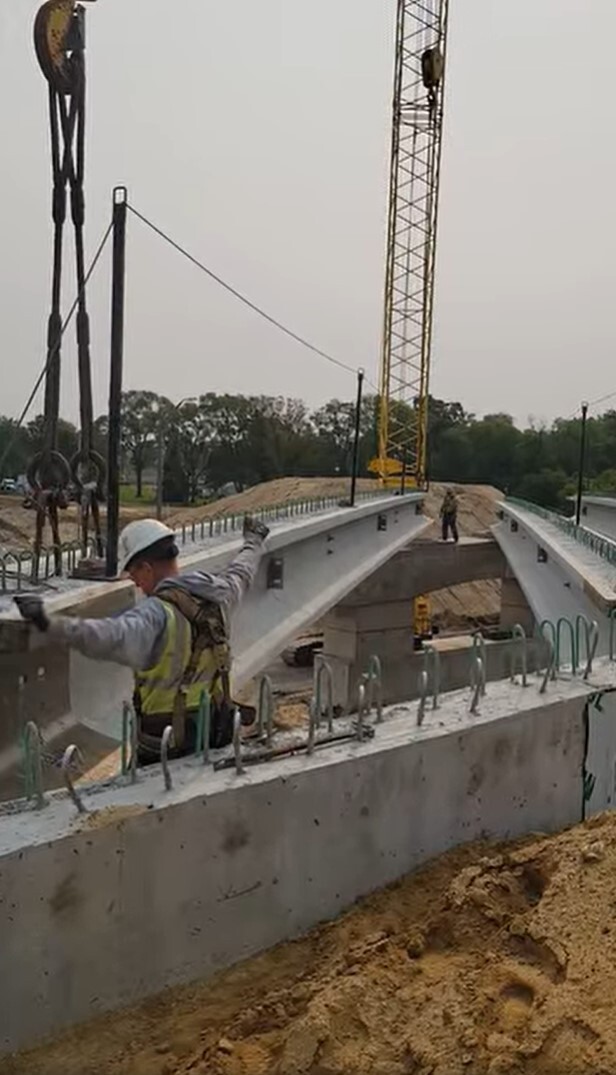 Bridge beam setting final placement with construction worker signaling placement.