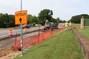 An orange detour sign, barrels, and safety fencing delineate a small construction zone on the side of a roadway.