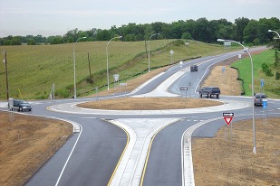 An elevated side view of a rural roundabout with fresh, brown sod shows three vehicles circulating and one exiting into the distance.