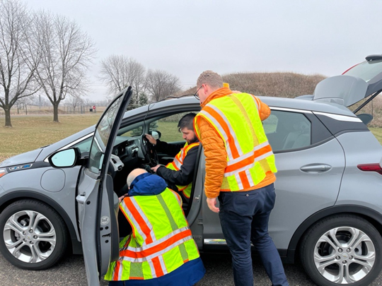 Three researchers with high visibility yellow vests inspect onboard data at the of the driver's footwell in a compact grey car.