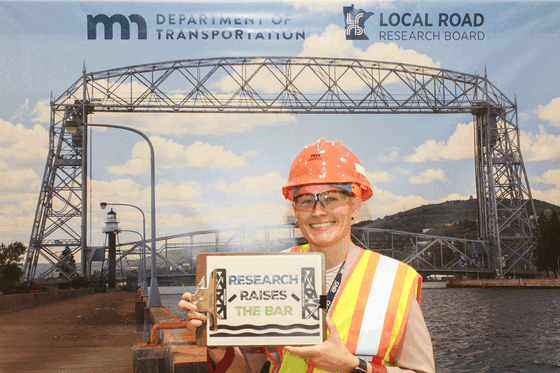 Animated GIF of people wearing hi vis PPE in front of Duluth's Aerial Lift Bridge photo banner holding a "Research Raises the Bar" sign