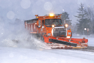 A bright orange MnDOT snowplow plows snow from a roadway during a snow event.