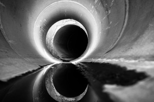 A black and white image from the interior of a round concrete culvert highlights joints and penetration by sunlight.