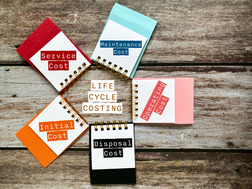 Small notebooks labeled with stages of LIfe Cycle Costing encircle a Life Cycle Costing label on top of weathered wood planks.