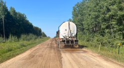 A large truck sprays liquid chemical stabilizer onto a gravel road from the rear of the vehicle.