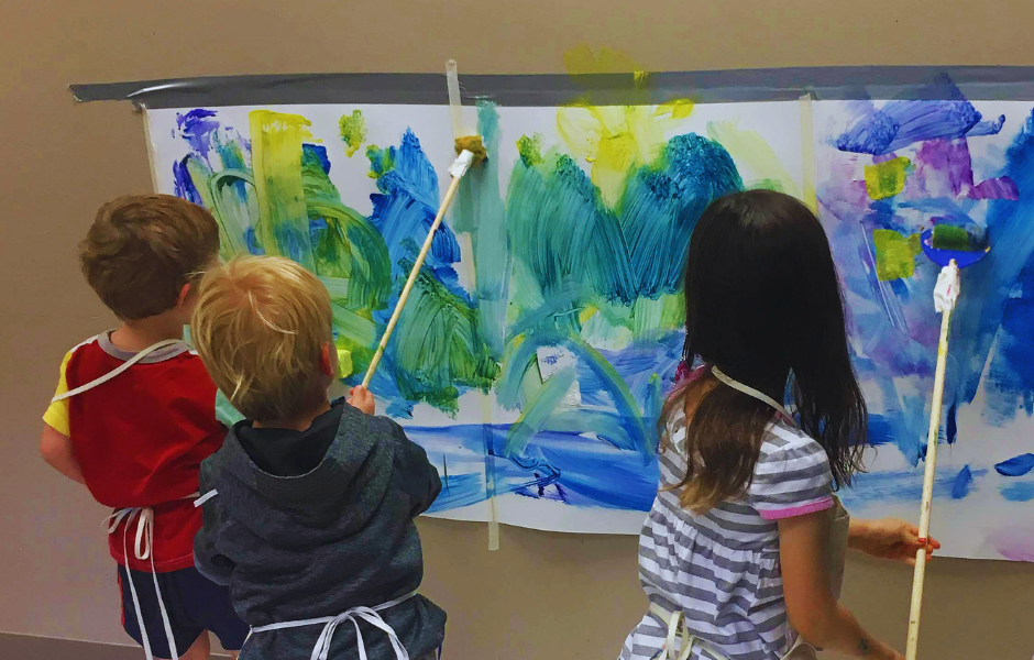 Three kids painting on a sheet of large paper hung up on wall.