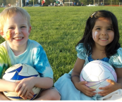Two young children sitting on a grassy field holding soccer balls