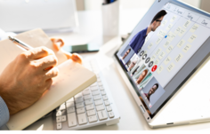 Person writing on a notebook while watching a presenter on the computer screen