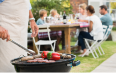 Man wearing apron while grilling meat and vegetables; people in background sitting at picnic table