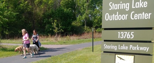 Two women walking their dogs on a paved path near the Staring Lake Outdoor Center Sign, 13765 Staring Lake Parkway