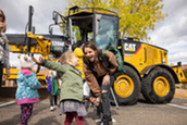 Woman laughing and looking toward a young girl as they stand in front of a yellow bulldozer