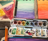 watercolor palette in a variety of colors, and three painted pieces in the background