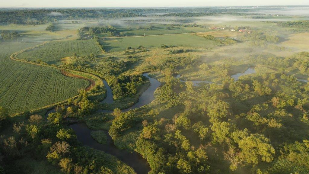 Aerial view of rural landscape with lush greenery, trees, river in the foreground and farmhouse, farmland in the background. 