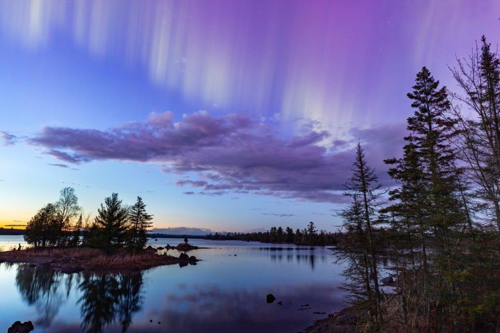 Northern lights erupt over a lake in Minnesota in a dark sky overhead shining rainbow of Aurora light as the sun sets on the horizon