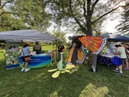 Pollinator event surrounded by trees, an attendee looks at the giveaways while a person in a butterfly suit looks at a flower model next to the table.