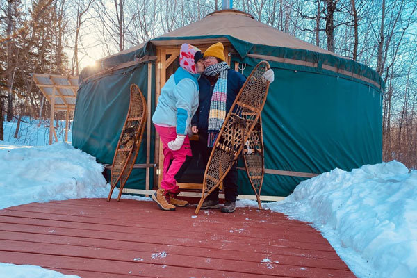 A couple kisses on the cheek and poses with snowshoes in front of their yurt