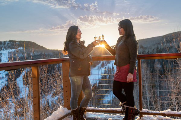 Two women toast drinks on an outdoor winter patio, with snow-covered mountains in background