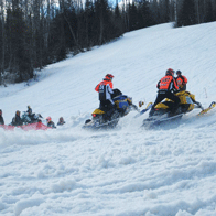 an image of snowmobilers racing up a hill with spectators in the background